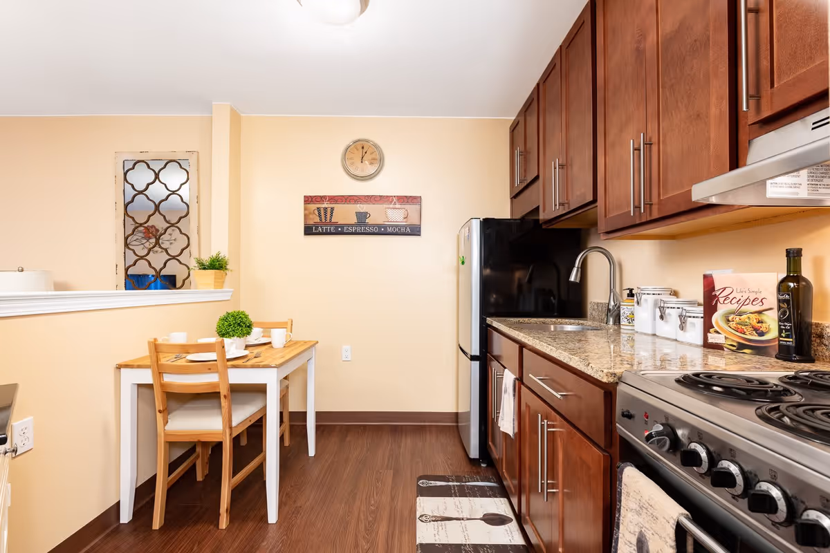 A small kitchen and dining area with wooden cabinets, a granite countertop, a stainless steel stove, and a black refrigerator. A wooden table with two chairs is set with white plates and cups. The wall has a clock and a decorative coffee-themed sign. The floor is dark wood, and the walls are painted light beige.
