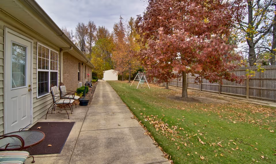 Outdoor walkway alongside a building with chairs and a small table on the left, leading to a white shed in the distance. On the right, there is a grassy area with scattered fallen leaves, a tree with red autumn foliage, and a swing set near a wooden fence.