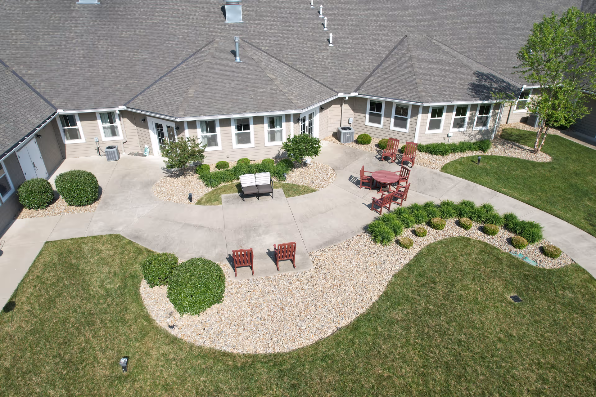 Aerial view of an outdoor patio area at Hudson Grande Senior Living featuring a concrete walkway, green grass, landscaped bushes, and several red wooden chairs around a round table and a bench.