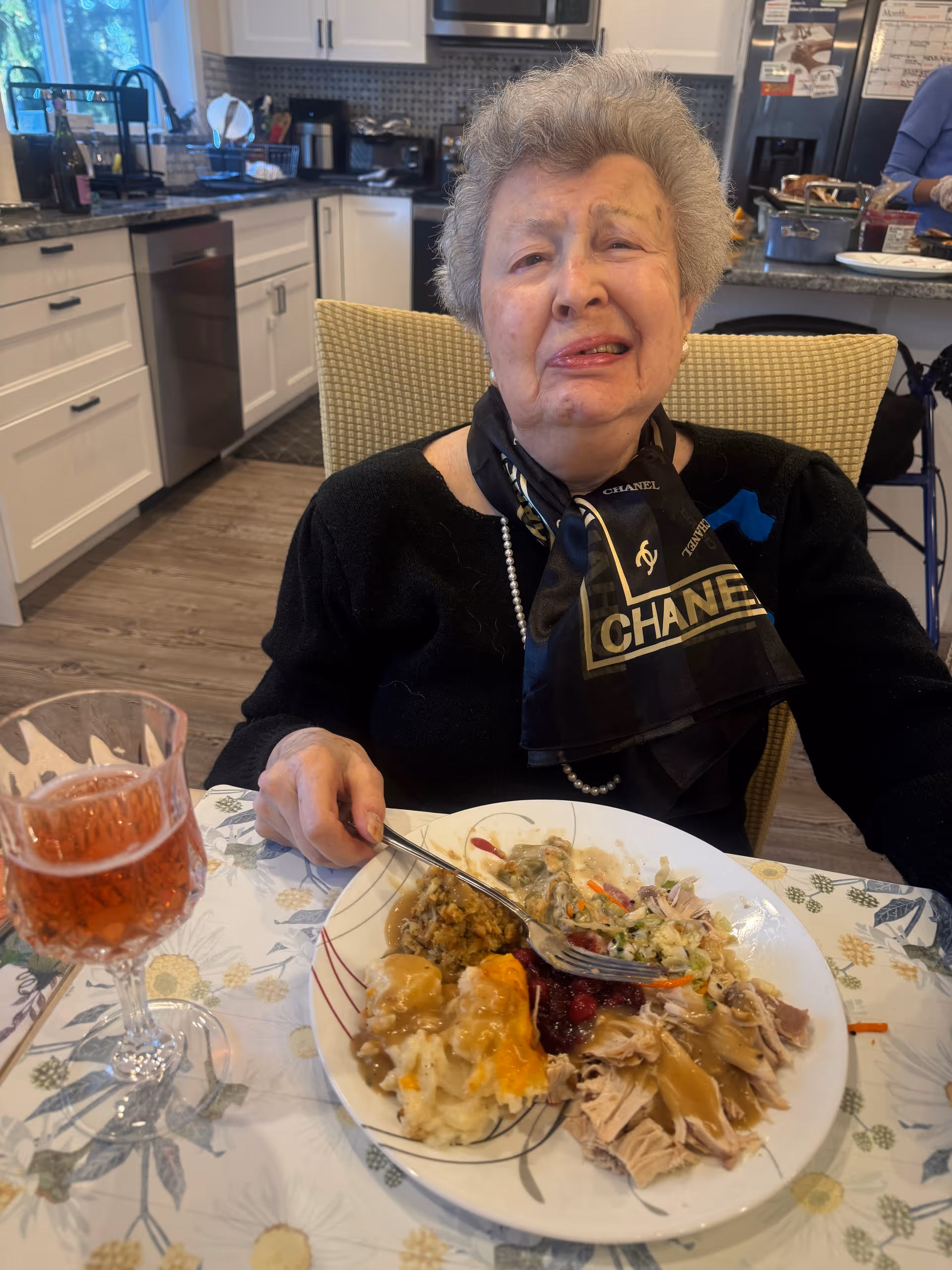 An elderly woman seated at a dining table with a plate of food and a glass of drink in a kitchen/dining area.
