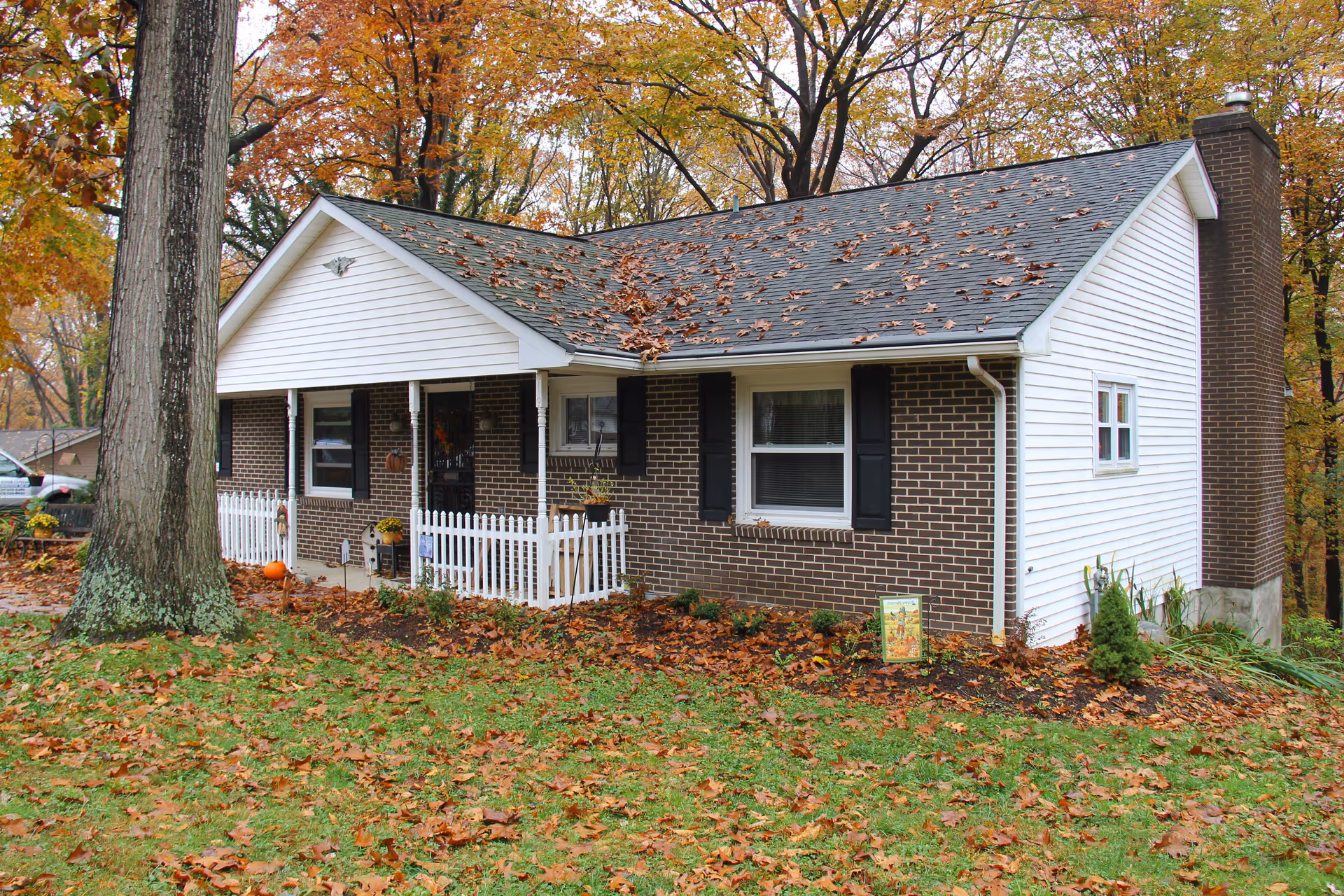 Exterior view of a single-story brick and white siding house with a small white picket fence in front, surrounded by autumn trees with fallen leaves on the roof and ground.