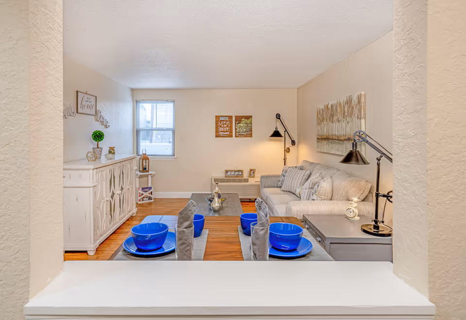 View through a pass-through window into a cozy living room with a beige sofa, decorative pillows, two black adjustable lamps, a wooden coffee table, and wall art. In the foreground, a dining table set with blue bowls, plates, and gray napkins is visible. The room has light-colored walls, a window with blinds, and a white sideboard with decorative items.