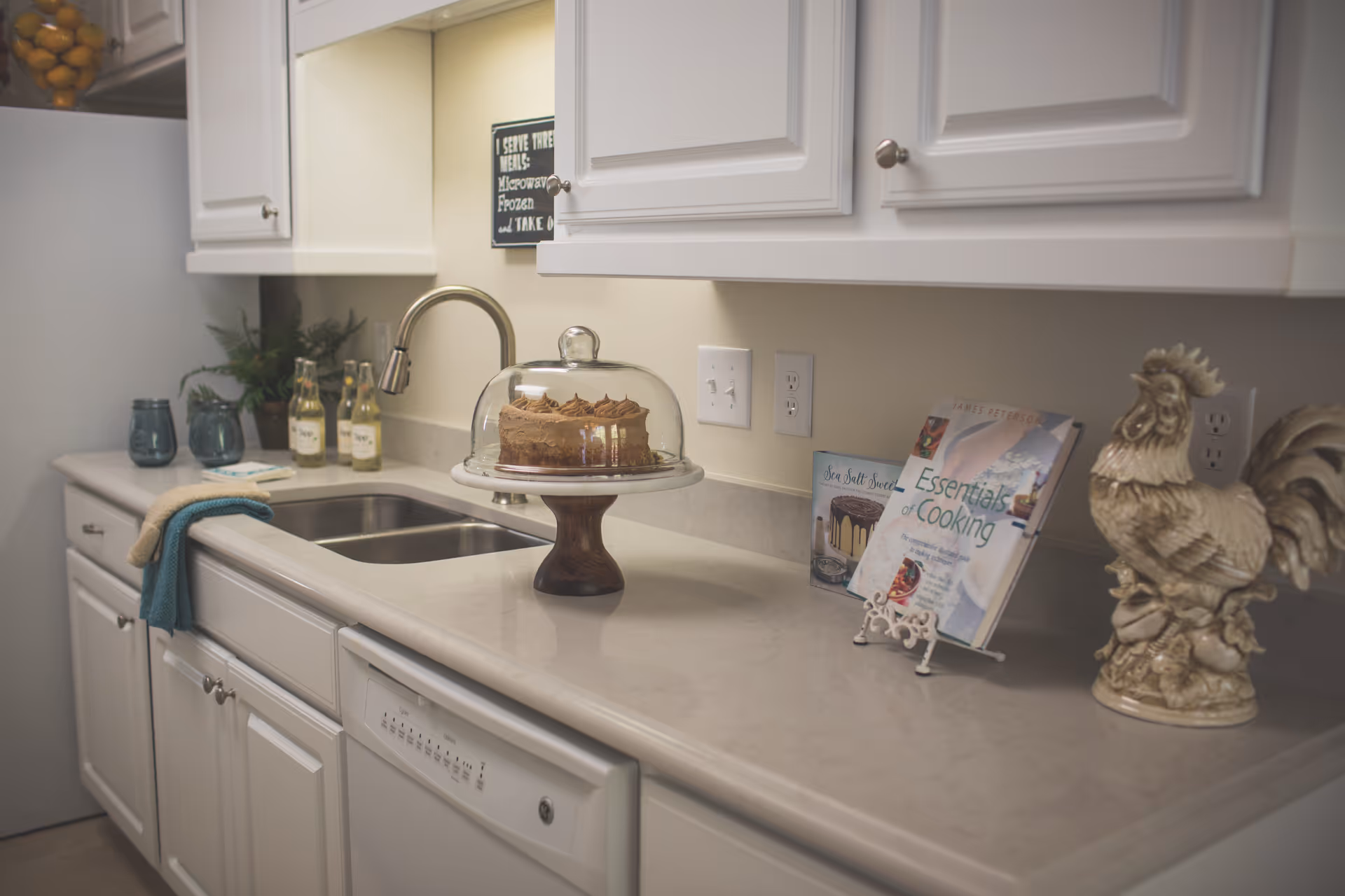 A clean kitchen countertop with a double sink, white cabinets, and a faucet. On the counter, there is a glass cake stand with a chocolate cake under a dome, two cookbooks on stands, a decorative ceramic rooster, two bottles of drink, two blue glasses, a small plant, and a towel hanging on the cabinet handle.