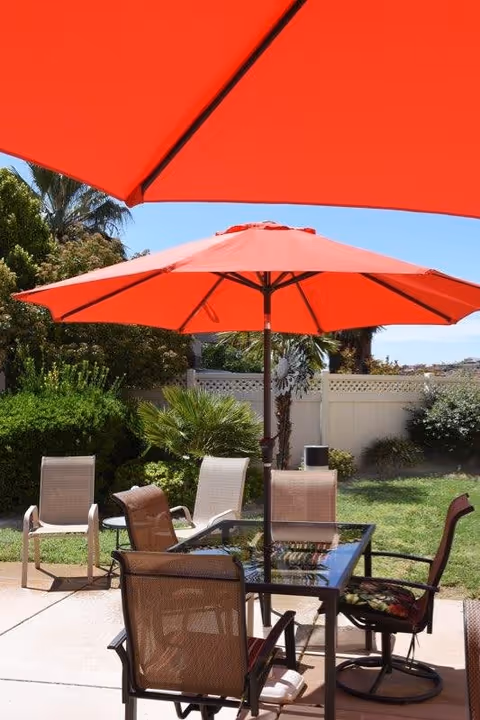 Outdoor patio area with a glass-top table surrounded by four mesh chairs, shaded by two large red umbrellas. The patio is adjacent to a grassy yard with various green shrubs and palm trees, enclosed by a white fence under a clear blue sky.