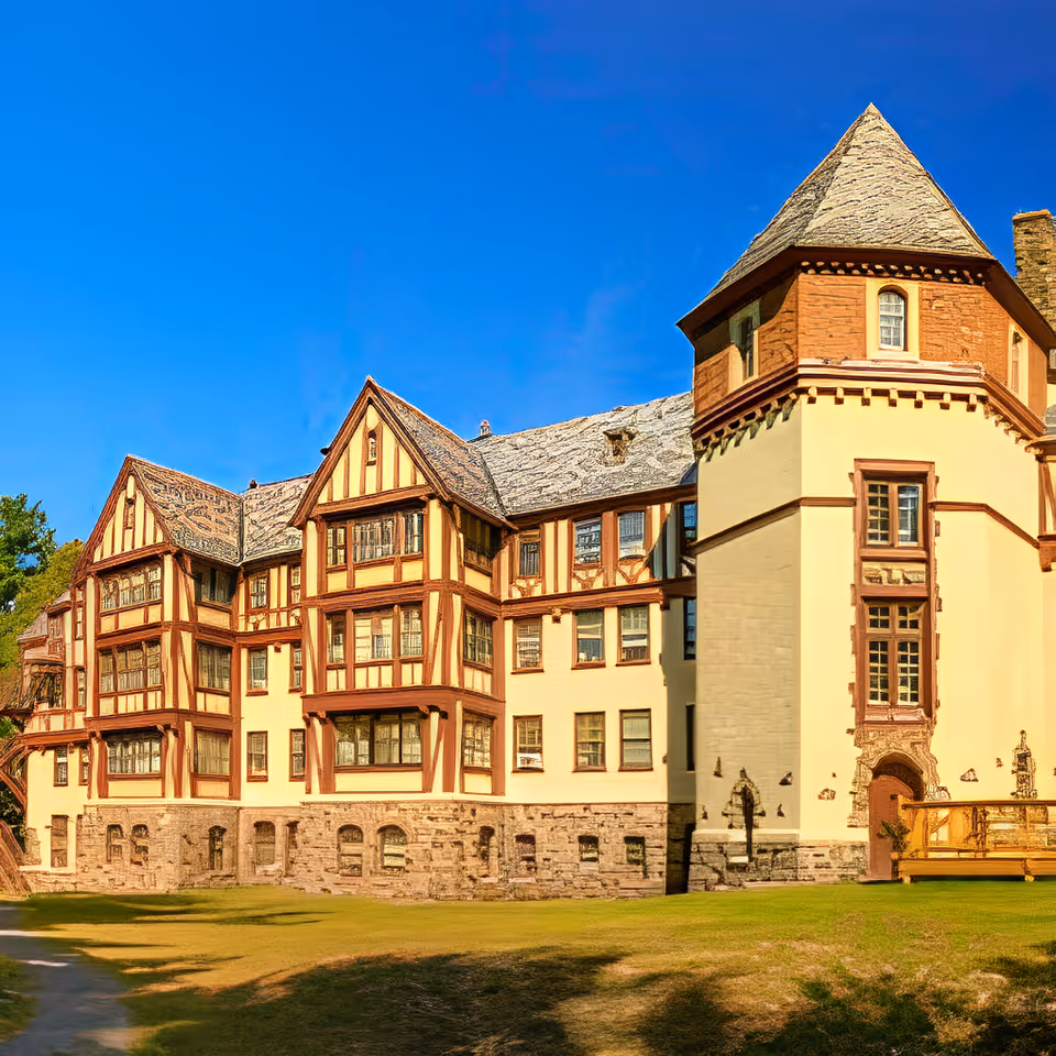 Exterior view of a large, historic-style building with multiple gabled roofs, timber framing, and stone foundation under a clear blue sky. The building has many windows and a prominent tower on the right side.