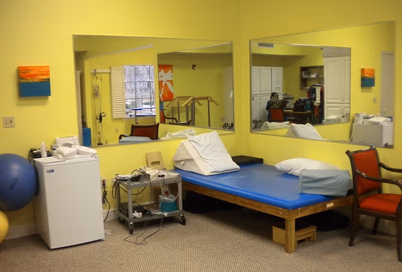 A therapy or treatment room with a blue padded treatment table, pillows, a small white refrigerator, exercise balls, a rolling cart with medical equipment, and two red chairs. The walls are painted yellow and have two large mirrors and colorful paintings. A window with white shutters is visible in the reflection.
