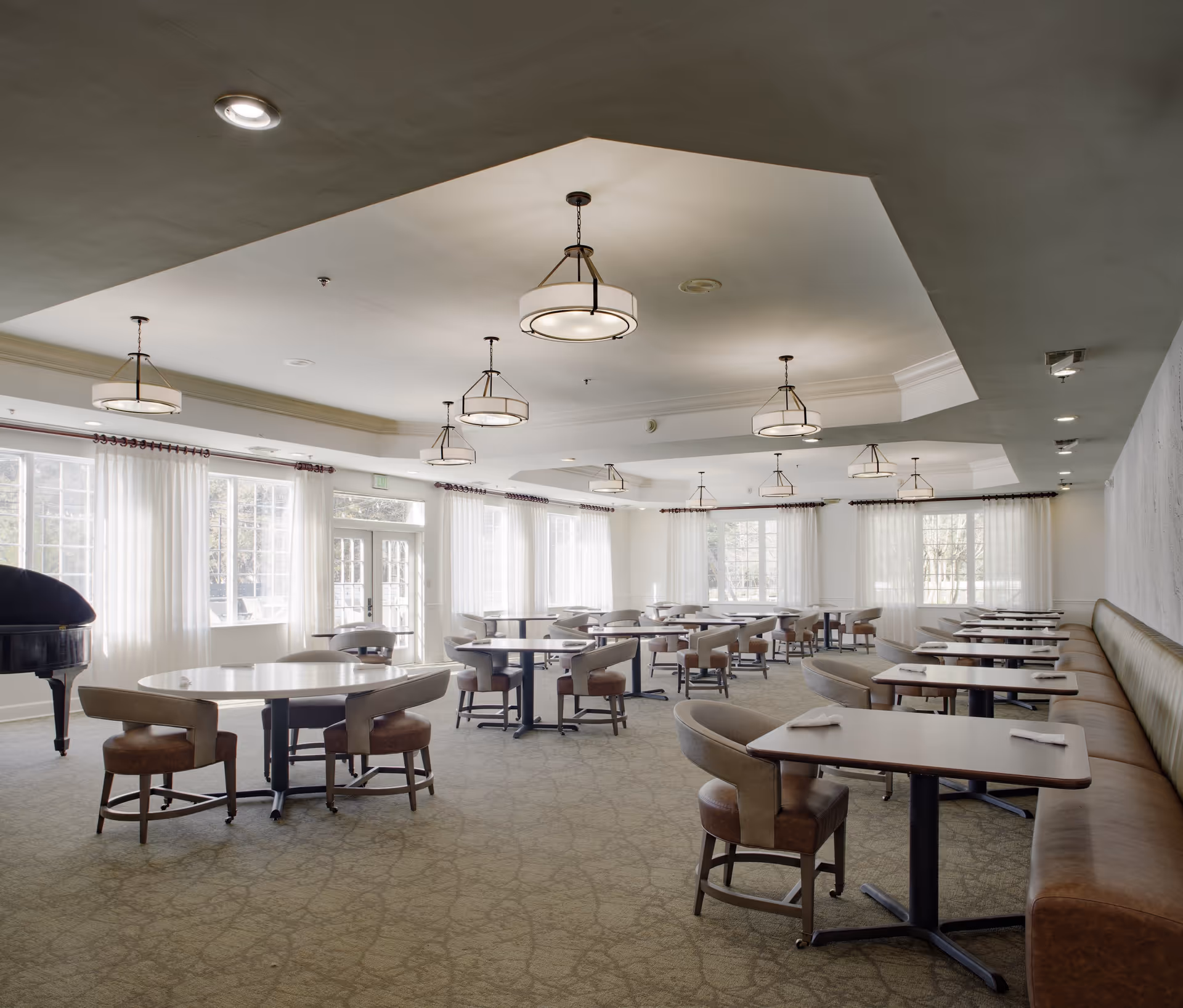 A spacious dining room with multiple tables and chairs arranged neatly. The room features large windows with sheer white curtains allowing natural light to fill the space. There are modern ceiling lights and a grand piano in the corner near the windows. The walls are light-colored, and there is a long cushioned bench along one side of the room.