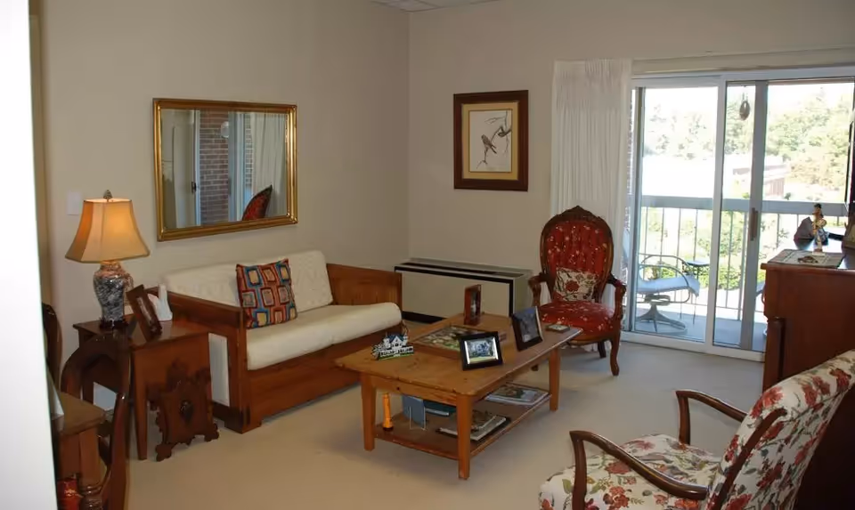 A cozy living room with a wooden framed sofa with white cushions and a colorful pillow, a wooden coffee table with picture frames and decorative items, two upholstered armchairs with floral and red patterns, a side table with a lamp, a framed bird artwork on the wall, a large mirror, and sliding glass doors leading to a balcony with outdoor seating and greenery visible outside.