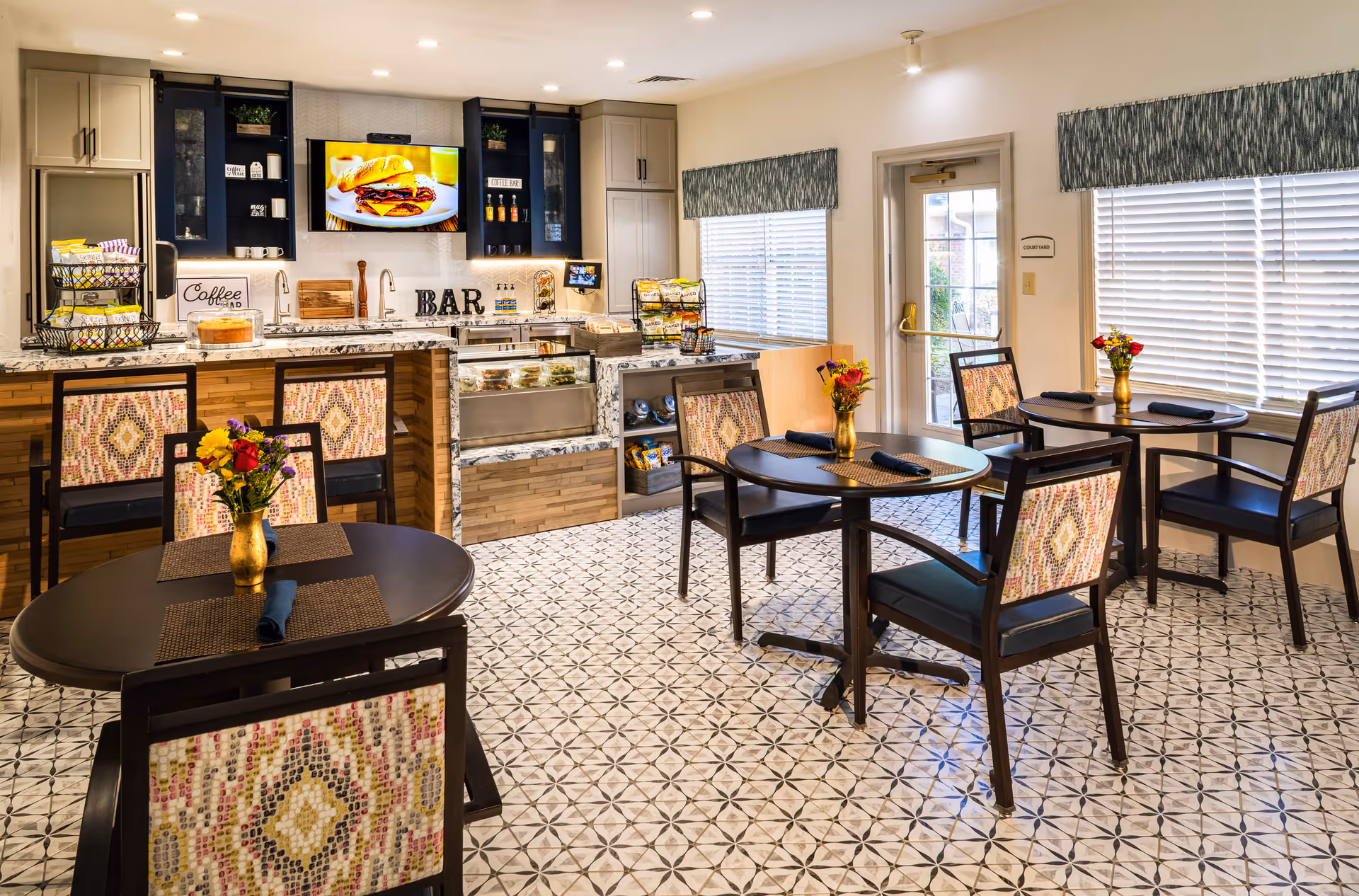 A bright and inviting dining area with several round tables and chairs featuring patterned upholstery. Each table has a small vase with colorful flowers and neatly folded napkins. In the background, there is a modern kitchen bar area with granite countertops, a coffee station, and a mounted TV displaying a sandwich. Large windows with blinds and valances let in natural light, and a door labeled 'Courtyard' leads outside.