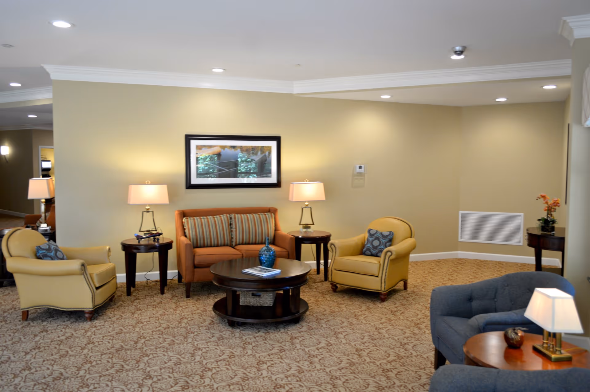 A cozy living room area in a senior living facility with beige walls and patterned carpet. The room features a brown sofa with striped cushions, two yellow armchairs with decorative pillows, a blue armchair, and three wooden side tables with lamps. A framed picture hangs on the wall above the sofa, and a small vase with flowers is on one of the side tables.
