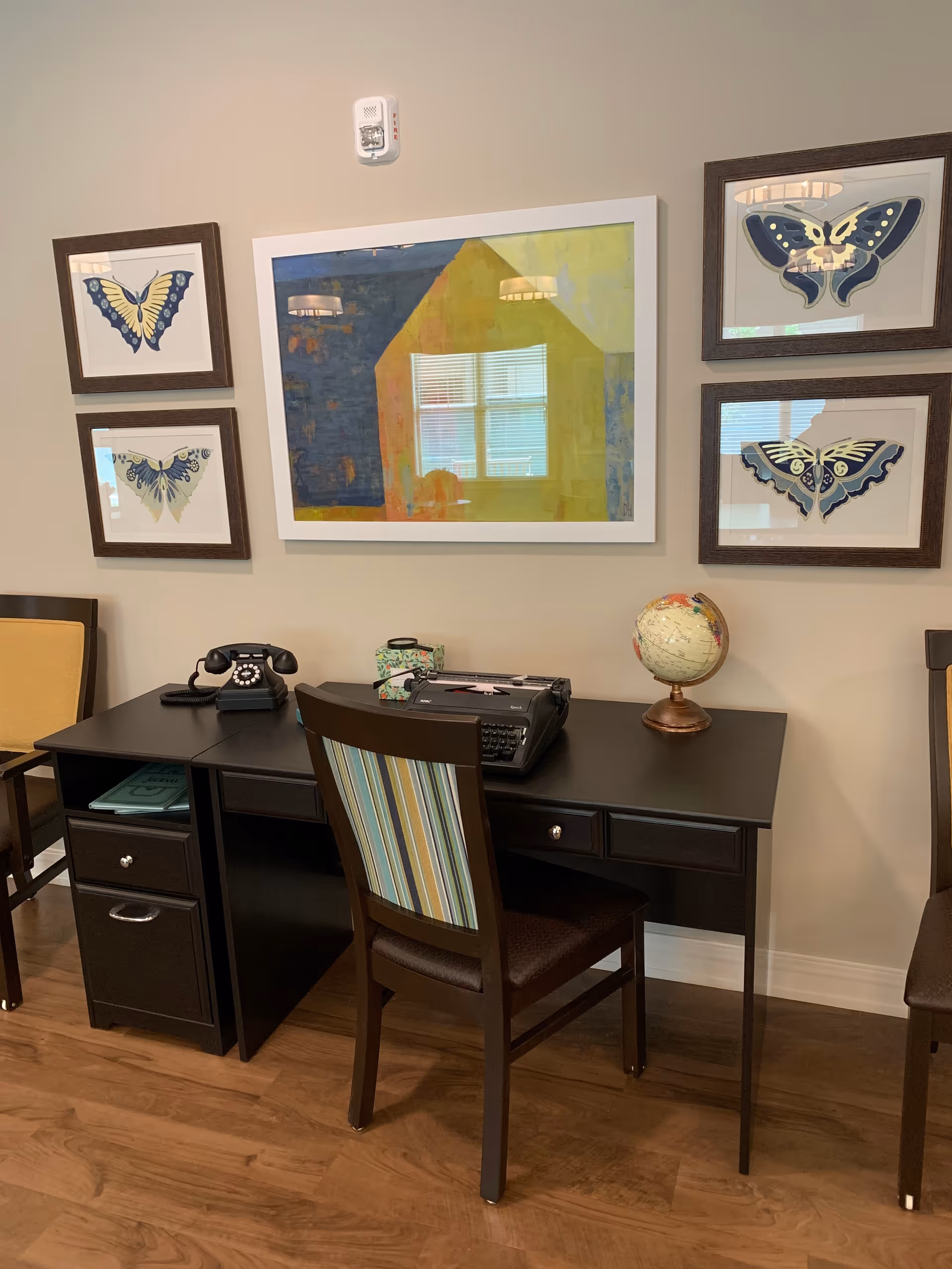A dark wooden desk with a vintage rotary phone, a typewriter, a small globe, and a decorative box on top. A chair with a striped backrest is placed in front of the desk. The wall behind the desk features five framed artworks, including four butterfly illustrations and one abstract painting. The floor is wooden.
