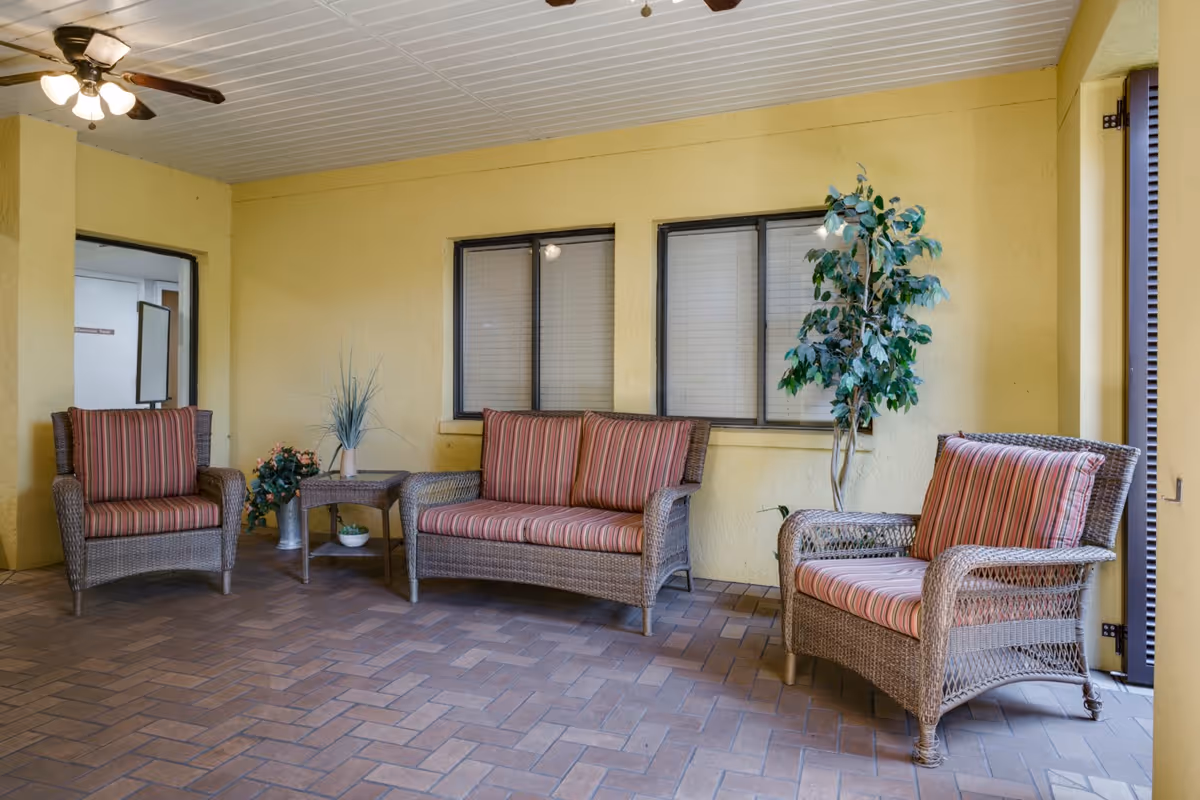 Covered seating area with wicker loveseat and two armchairs with striped cushions, a small side table, potted plants, and a tiled floor.