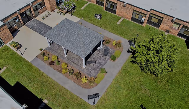 Aerial view of an outdoor courtyard area at Alpine Rehabilitation & Nursing Center featuring a covered pavilion with a shingled roof, surrounded by paved walkways, green grass, shrubs, a tree, and benches. The courtyard is enclosed by a brick building with multiple windows and doors.