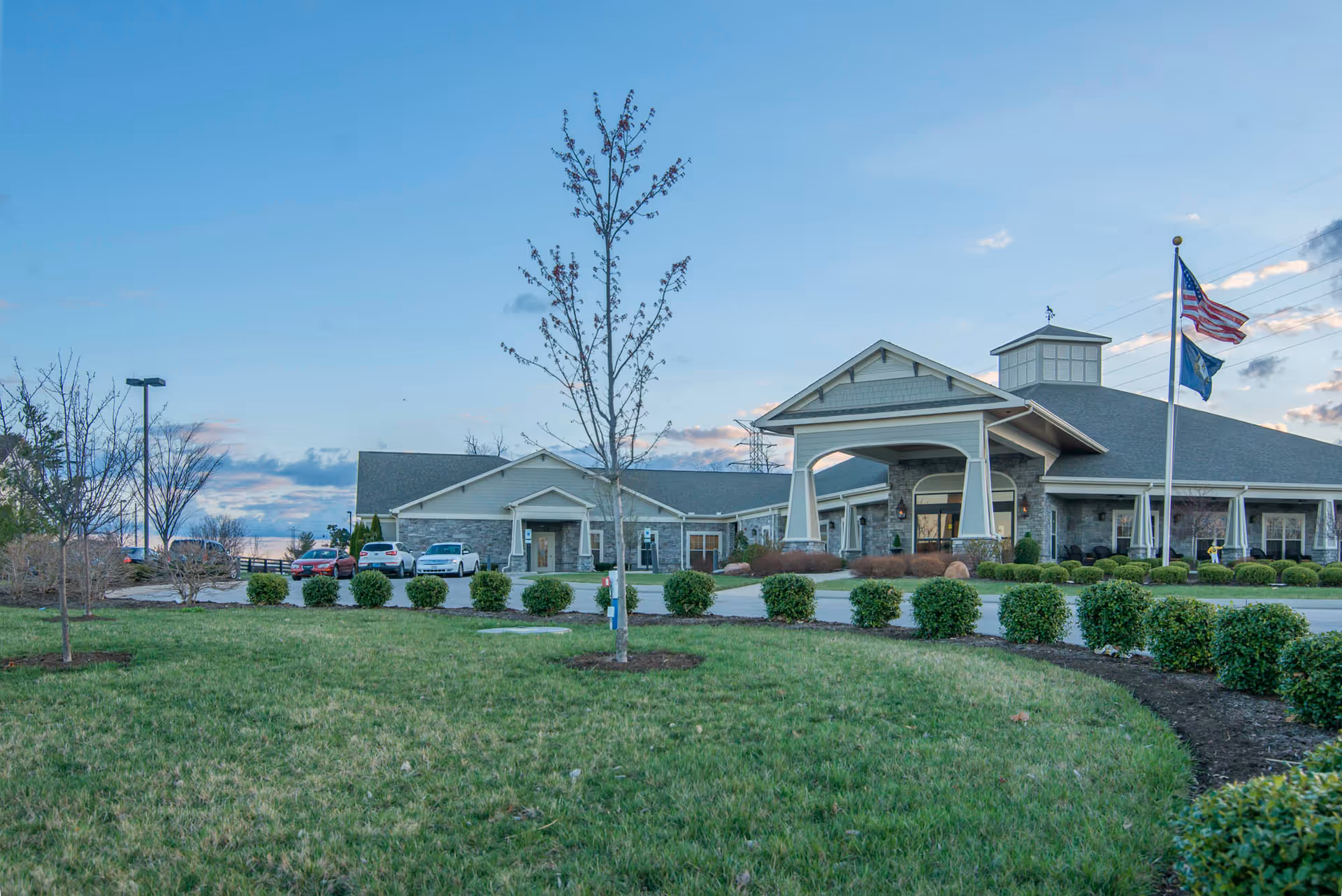 Exterior view of Morning Pointe of Lexington facility with a manicured lawn, small trees, bushes, a driveway with parked cars, and two flagpoles displaying the American flag and another flag under a partly cloudy sky.