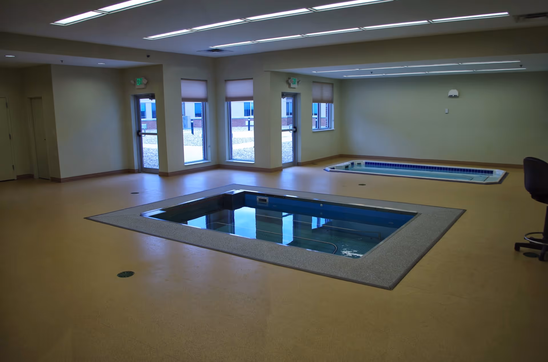 Interior therapy room with two recessed small pools/spa tubs, windows and doors, and fluorescent ceiling lights.