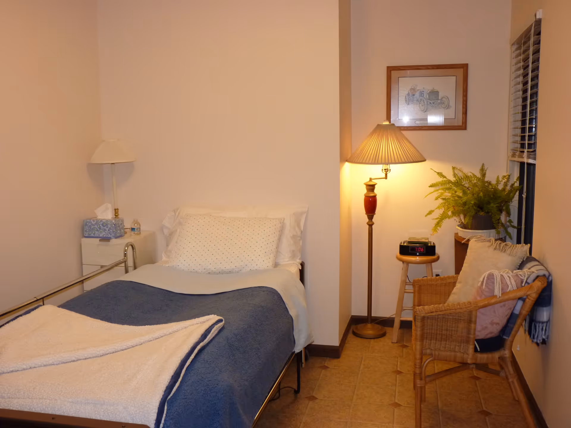 A small, cozy bedroom with a single bed covered with a blue blanket and a white throw. There is a white pillow with small dots and a white head pillow. Next to the bed is a small white nightstand with a lamp, tissue box, and water bottle. On the right side, there is a wooden chair with cushions and a blanket, a small wooden stool with a digital clock showing 7:06, a floor lamp with a pleated shade, a green potted plant, and a framed picture on the wall. The room has tiled flooring and a window with blinds.