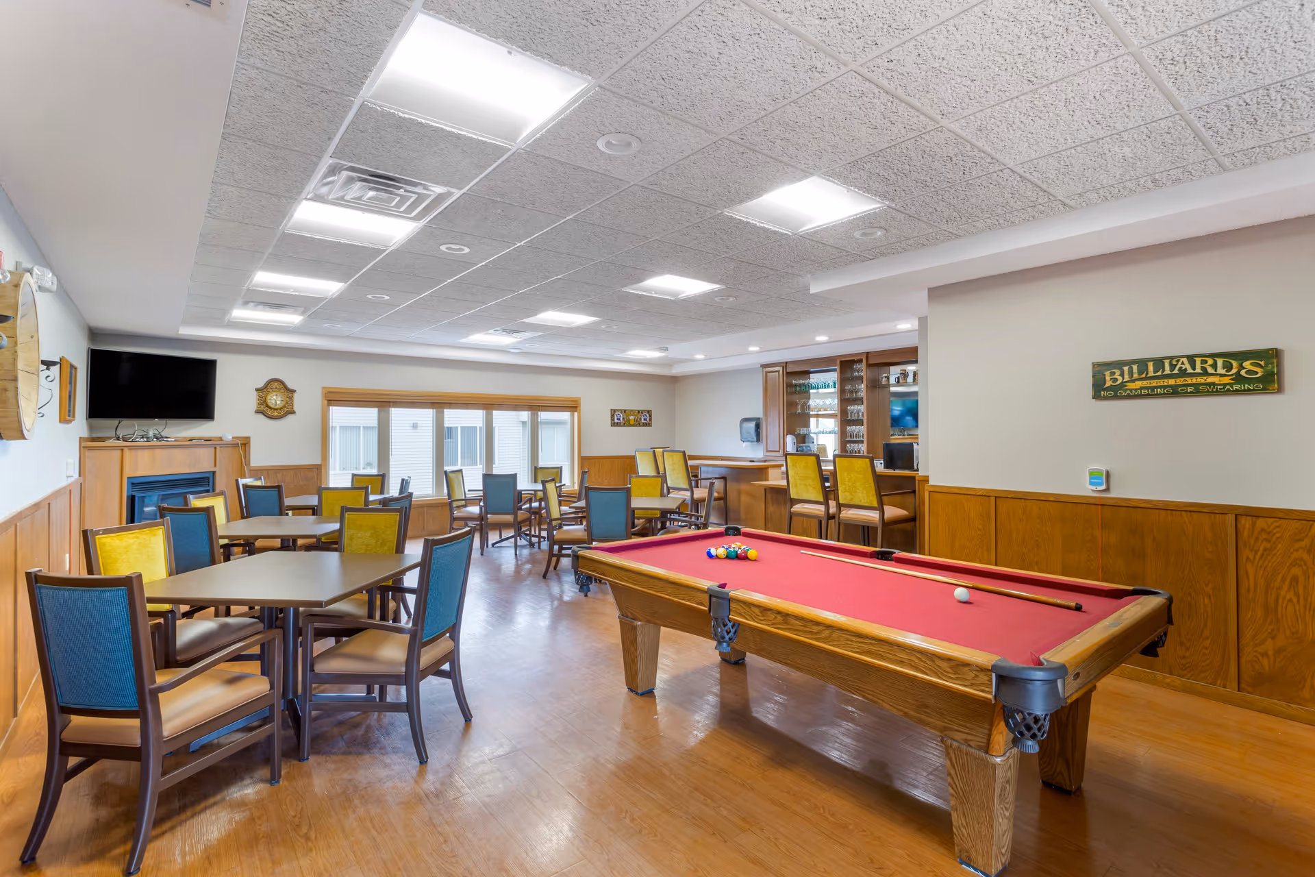 A senior living facility common room featuring a red felt pool table with billiard balls and cues, several tables and chairs with blue and yellow upholstery, a wall-mounted TV above a fireplace, and a bar area with high chairs and glassware. The room has wood paneling on the lower walls and large windows letting in natural light.