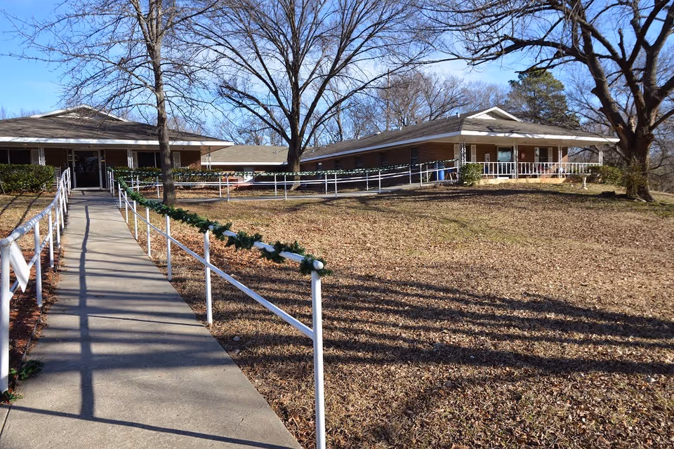 Single-story nursing facility with a paved walkway and white handrails leading to the front entrance, set on a lawn with bare trees.
