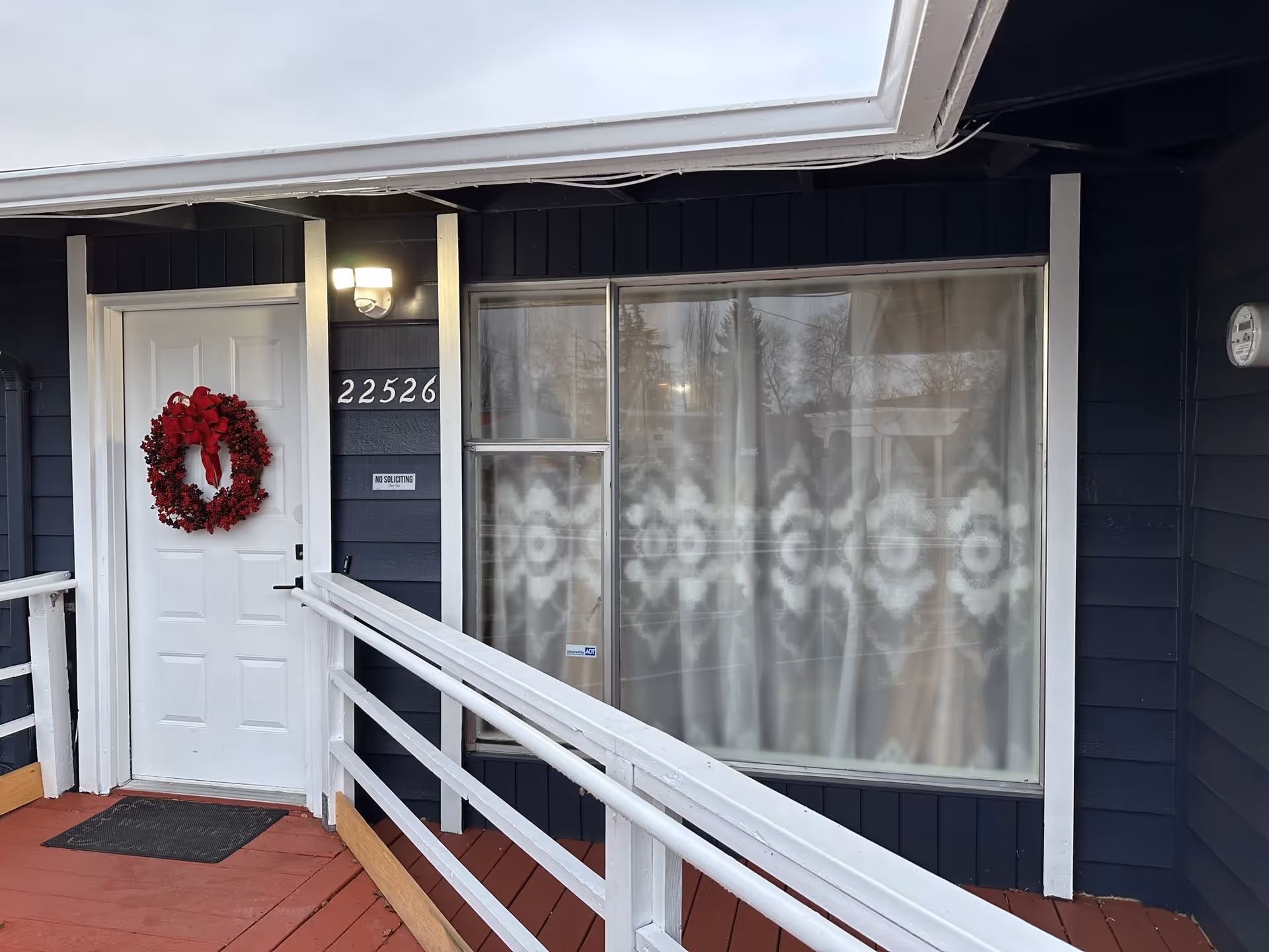 Entrance of a building with a white door decorated with a red wreath, a window with patterned curtains, a white railing along a red wooden ramp, and the address number 22526 displayed on the dark blue exterior wall.