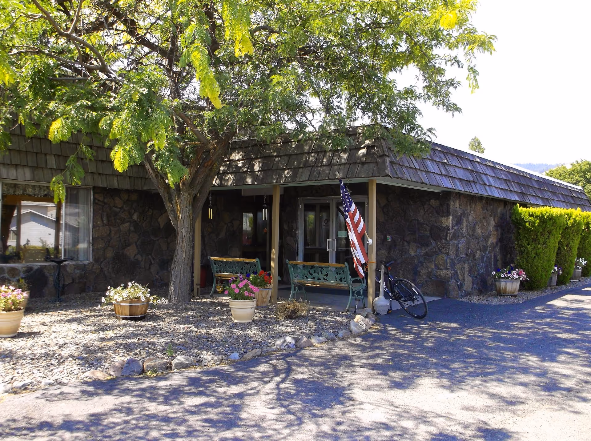 Exterior view of a stone building with a shingled roof, featuring a covered entrance with two green benches and an American flag. There are several potted plants with flowers around the entrance and a bicycle leaning against one of the benches. A large tree with green leaves provides shade in front of the building.