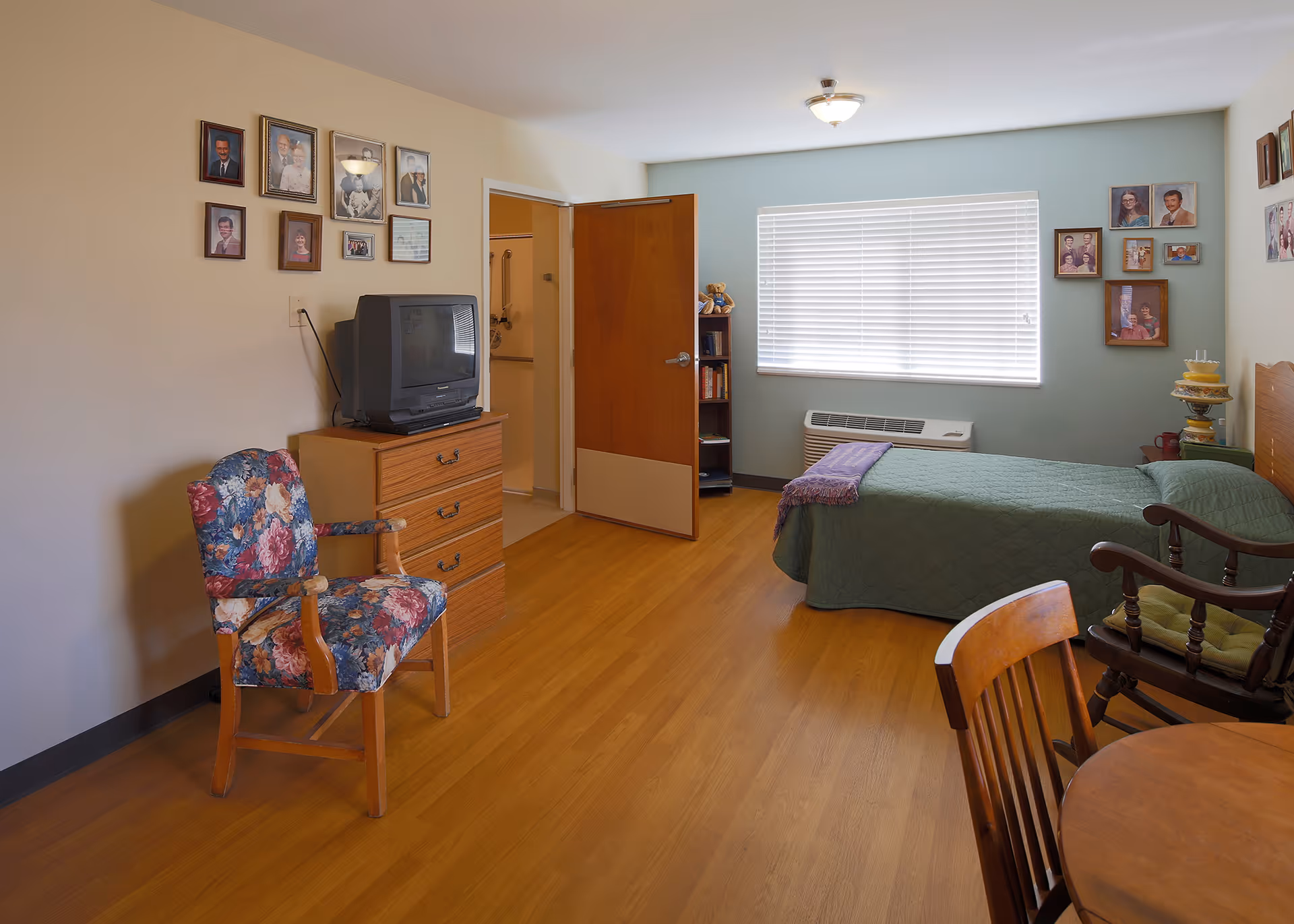 Furnished memory-care bedroom with a single bed, dresser and TV, floral armchair, dining table chairs, and family photos on the walls.