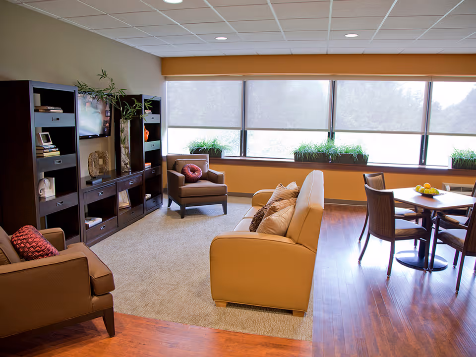 A bright and cozy living room area with a beige sofa and two brown armchairs, each with decorative pillows. There is a dark wooden entertainment center with shelves holding books and decorative items, and a flat-screen TV mounted on the wall. Large windows with roller shades let in natural light, and there are green plants on the windowsill. A small dining table with four chairs and a bowl of fruit is positioned on the wooden floor near the windows.