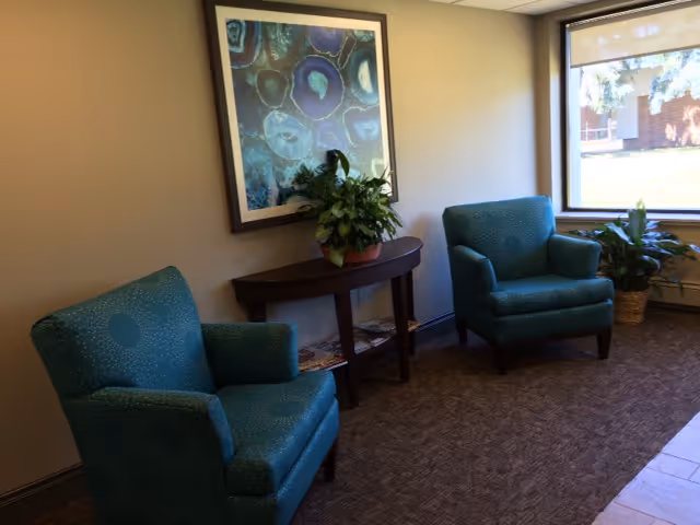 A cozy sitting area with two teal upholstered armchairs placed on a brown carpeted floor. Between the chairs is a dark wooden console table with a potted plant on top. Above the table hangs a framed abstract artwork featuring blue and green circular patterns. A large window on the right side lets in natural light and shows a view of greenery outside. Another potted plant is positioned near the window.
