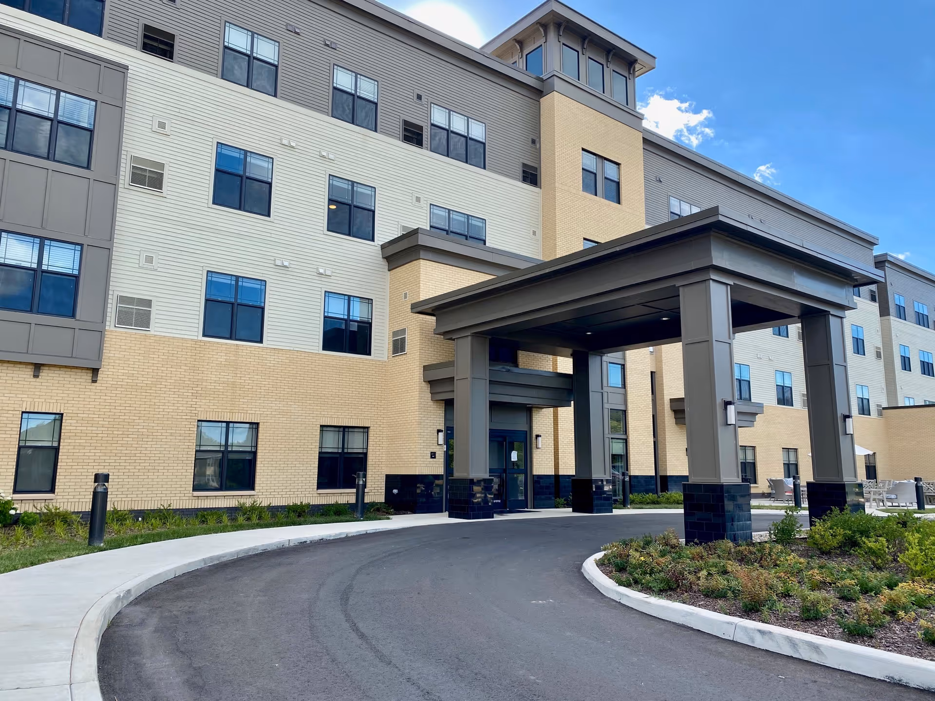 Exterior view of a multi-story senior living facility building with a covered entrance driveway, landscaped greenery, and clear blue sky.