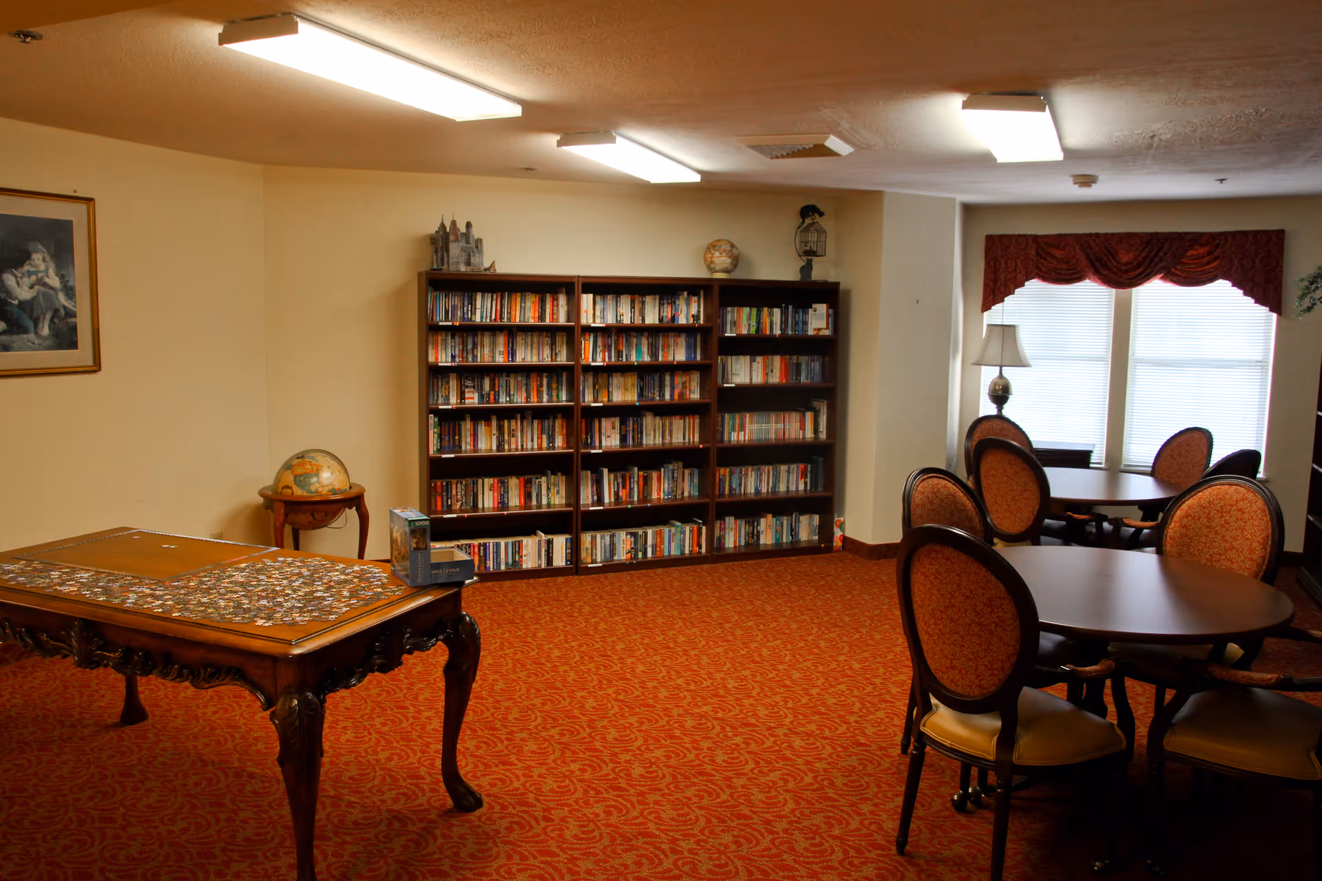 A cozy room with red patterned carpet, featuring a wooden table with a puzzle in progress, a globe on a small side table, a large bookshelf filled with books, round tables with upholstered chairs, a floor lamp, and windows with red valances letting in natural light.