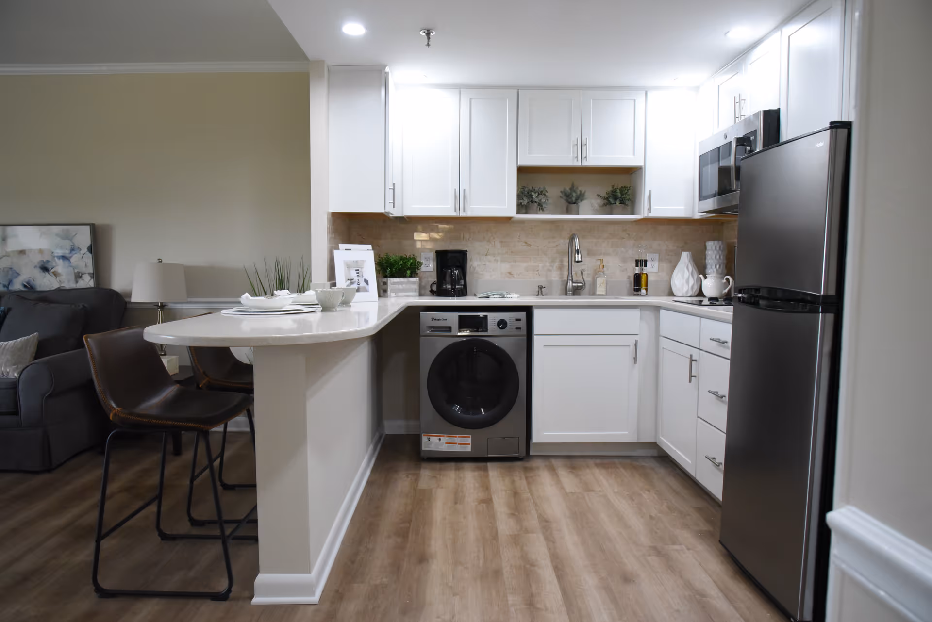 Modern kitchen area with white cabinets, a stainless steel refrigerator, microwave, and a washing machine under the counter. There is a countertop with two brown bar stools and a small dining setup with plates and cups. The kitchen opens into a living area with a gray sofa and a lamp.