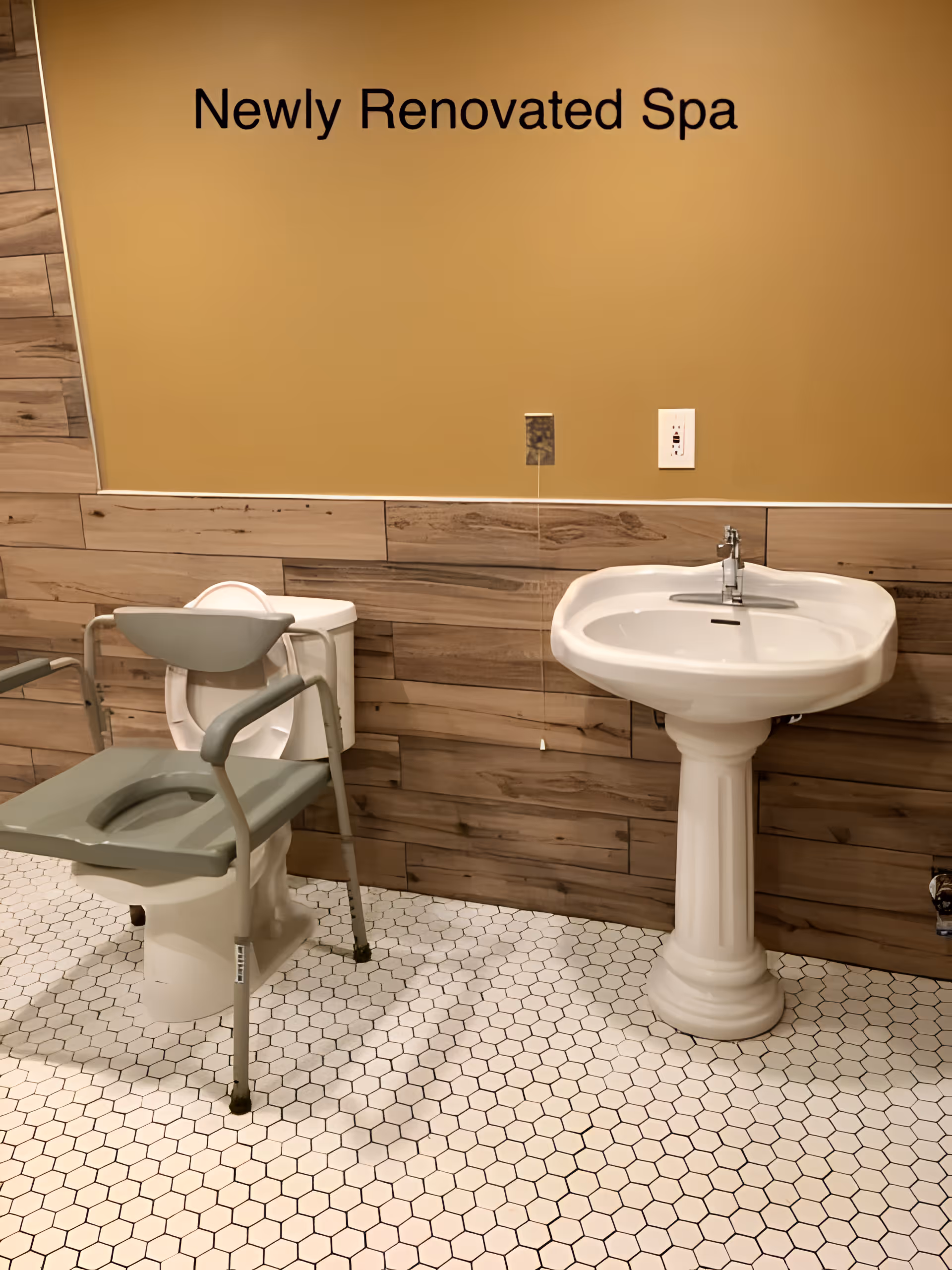 Interior view of a newly renovated spa bathroom featuring a white pedestal sink and a toilet with a gray raised toilet seat and armrests. The walls have wood paneling halfway up and are painted a warm beige color above. The floor is covered with white hexagonal tiles. A sign on the wall reads 'Newly Renovated Spa'.