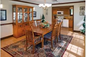 Wooden dining table with six chairs on a patterned rug in a bright dining area with a china cabinet and a pass-through to the kitchen.