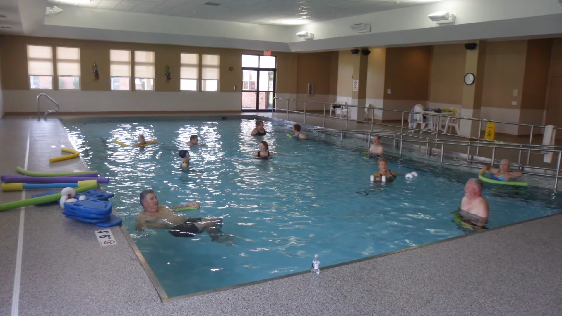 Indoor swimming pool with several elderly people in the water using pool noodles and flotation devices. The pool area has large windows with blinds, beige walls, and a handrail along one side. Pool equipment is stacked on the pool deck.