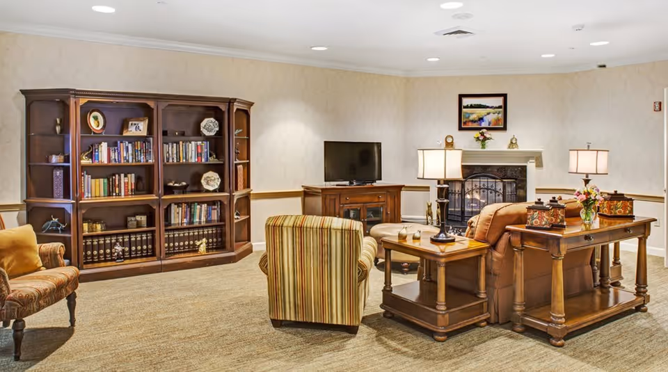 A cozy living room in a senior living facility featuring a wooden bookshelf filled with books and decorative items, a TV on a wooden stand, a fireplace with a painting above it, and comfortable seating including a striped armchair and a brown sofa with side tables and lamps.