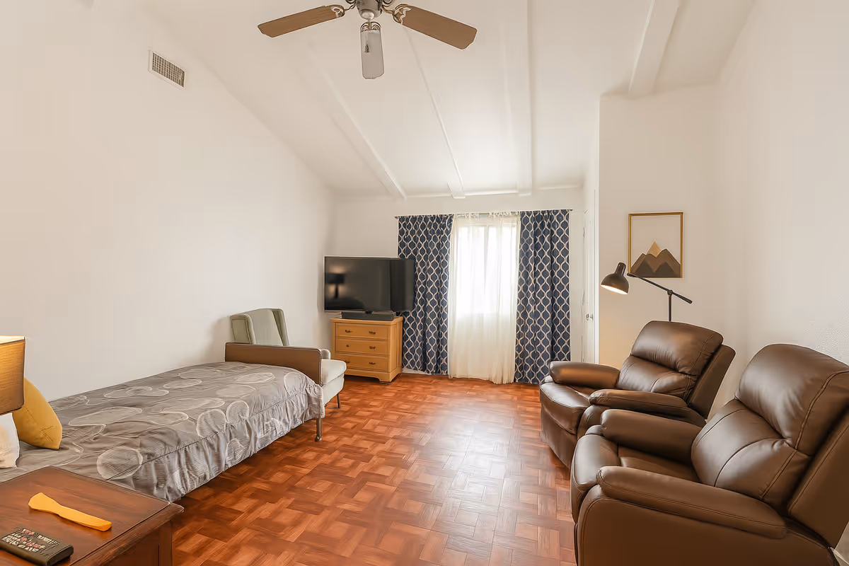 A well-lit room with a single bed covered in a gray patterned blanket, a green armchair, a wooden dresser with a flat-screen TV on top, two brown leather recliners, a floor lamp, and a window with blue and white patterned curtains letting in natural light.