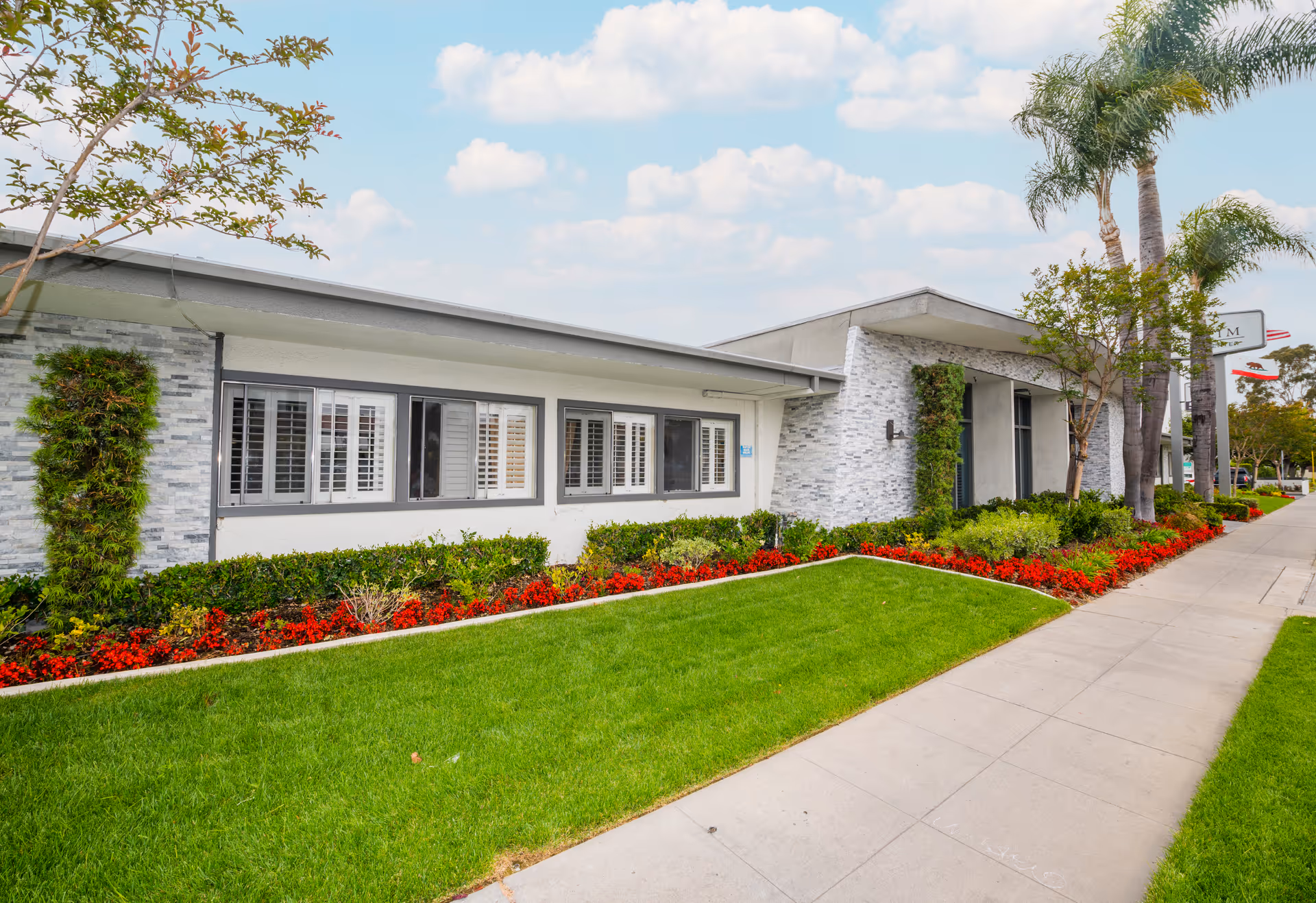 Exterior view of Anaheim Healthcare Center showing a single-story building with white and gray stone walls, several windows with white shutters, well-maintained green lawn, red flowers, shrubs, and palm trees along a sidewalk under a partly cloudy sky.
