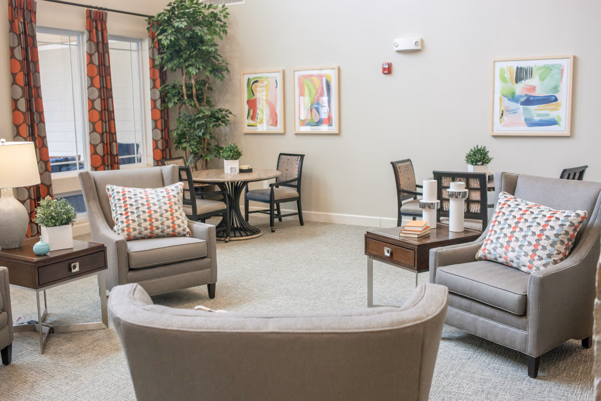 A cozy living room area with three gray armchairs, each with a patterned cushion. There are two wooden side tables with decorative items including a lamp, small plants, and candles. In the background, there are two round tables with chairs, colorful abstract paintings on the wall, and a tall green plant near the window with red and gray patterned curtains.