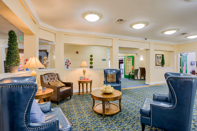 A cozy senior living facility lounge area with blue leather armchairs and a brown leather chair arranged around a round wooden coffee table. The room has patterned carpet, cream-colored walls, decorative plants, table lamps, and framed artwork. There are open wall sections and doorways leading to other rooms.