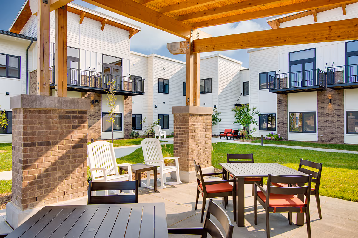 Outdoor patio area at Preston Greens Senior Living with wooden pergola supported by brick pillars, several tables and chairs with red cushions, white plastic chairs, green grass, and a modern two-story building with balconies in the background under a partly cloudy sky.