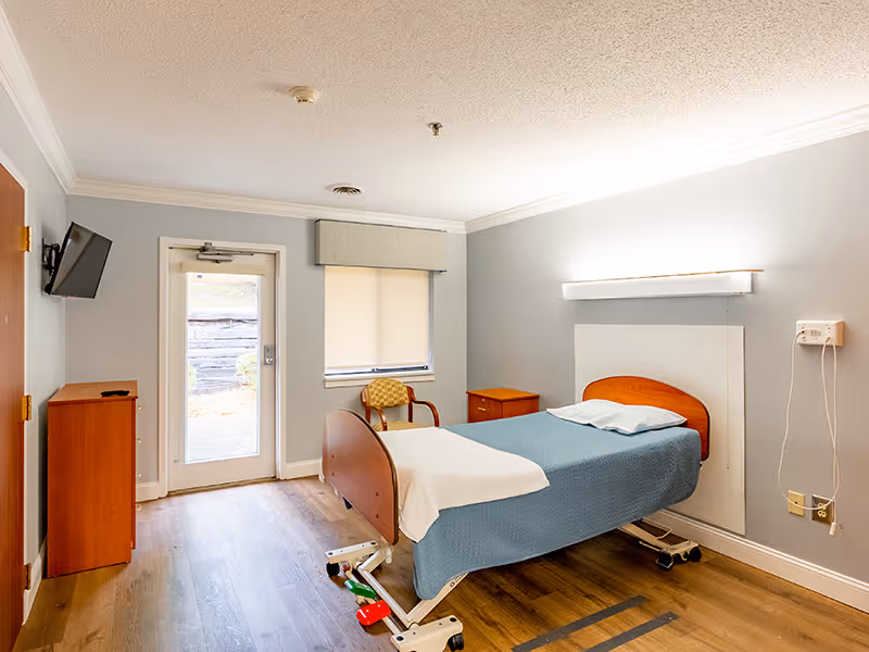 A simple, clean bedroom in a senior living facility with a single adjustable bed covered with a blue blanket and white sheet. The room has light gray walls, a wooden floor, a small wooden nightstand, a chair with a patterned cushion, a wall-mounted TV, and a door with a window leading outside.