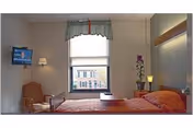 A small, neatly arranged bedroom with a single bed covered in an orange bedspread. There is a wooden chair with an orange cushion next to the bed, a wall-mounted television, a window with a valance, and a small table with a lamp and a potted orchid.