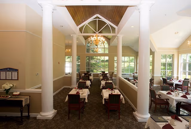 Dining room with multiple tables covered in white tablecloths and set with cups and napkins. The room features large windows letting in natural light, white columns, chandeliers, and a high ceiling with wooden accents.