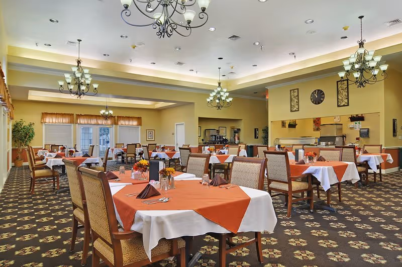 A spacious dining room with multiple tables covered in white and orange tablecloths, each set with glasses, napkins, and utensils. The room features patterned carpet, chandeliers hanging from the ceiling, and a clock on the wall above a serving area. Large windows with curtains allow natural light to enter the room.