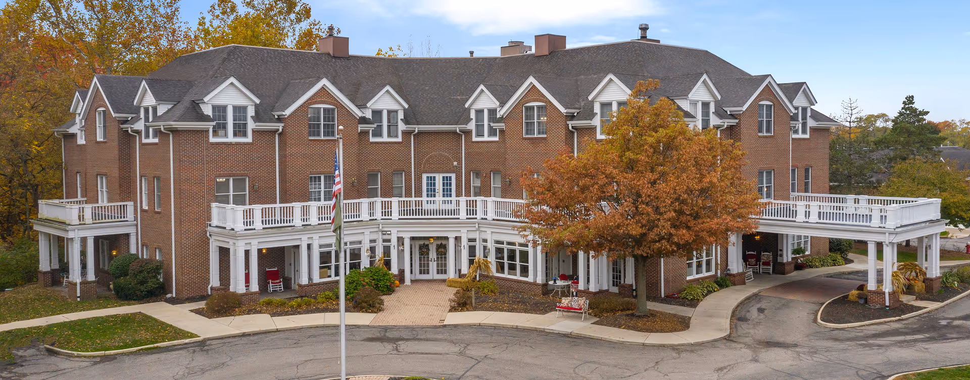 Front exterior view of a large brick senior living facility with white trim and multiple windows. The building has a covered entrance with white columns and a circular driveway. There is an American flag on a flagpole in front, and a tree with autumn-colored leaves near the entrance.