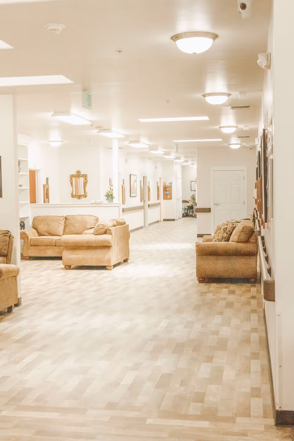 A well-lit hallway in a senior living facility with beige couches along the walls, framed pictures, and a mirror. The floor has a light wood pattern, and the ceiling has multiple light fixtures. There is a person in a wheelchair visible in the distance.