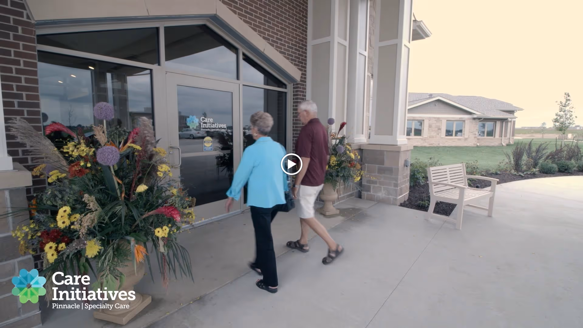 Two elderly people walking towards the entrance door of a building with large flower arrangements on either side of the door. The building has brick and stone exterior with a bench nearby and a grassy area in the background.