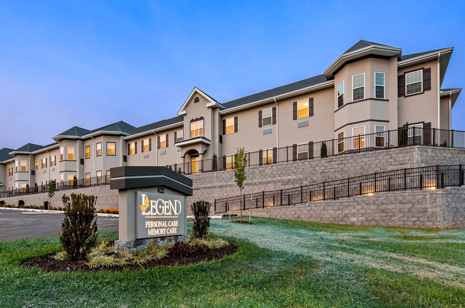 Exterior view of a large, multi-story senior living facility building at dusk with lights on inside. In the foreground, there is a landscaped area with a sign that reads 'Legend Personal Care Memory Care' and the number 425. The building has multiple windows and a sloped roof.