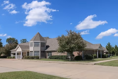 Exterior view of a single-story senior living facility building with a turret-like architectural feature, surrounded by green lawns and trees under a blue sky with scattered clouds.