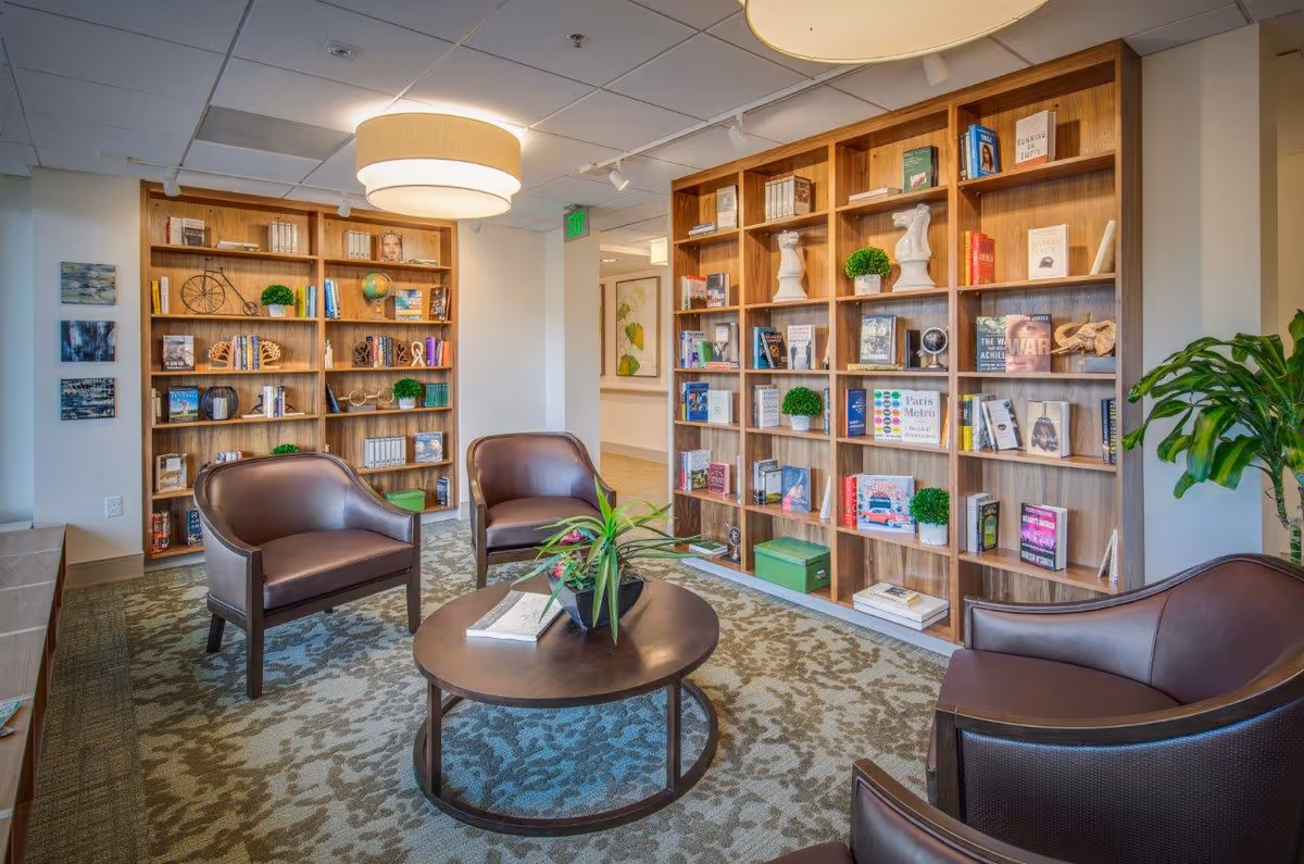 A cozy reading or sitting area in a senior living facility featuring wooden bookshelves filled with books and decorative items, four brown leather armchairs arranged around a round wooden coffee table with a plant and books on it, a patterned carpet, and a modern ceiling light fixture.