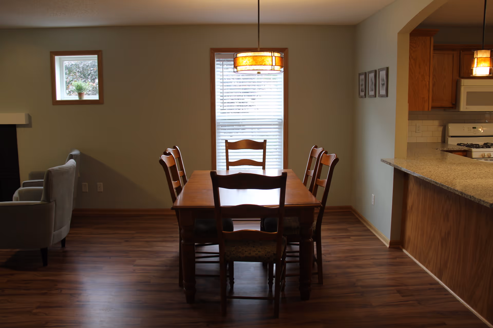 A dining area with a wooden table and six matching chairs on a wood floor. A hanging light fixture is centered above the table. To the right is a kitchen counter with a granite countertop and wooden cabinets. In the background, there is a window with blinds and a small square window with a potted plant. Two gray armchairs are partially visible on the left side of the image.